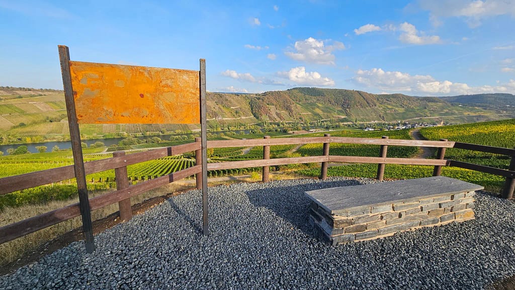 Steinbank und Geländer am Sitzplatz der Bienendisko auf dem Galgisberg mit Blick auf Weinberge und Mosel.