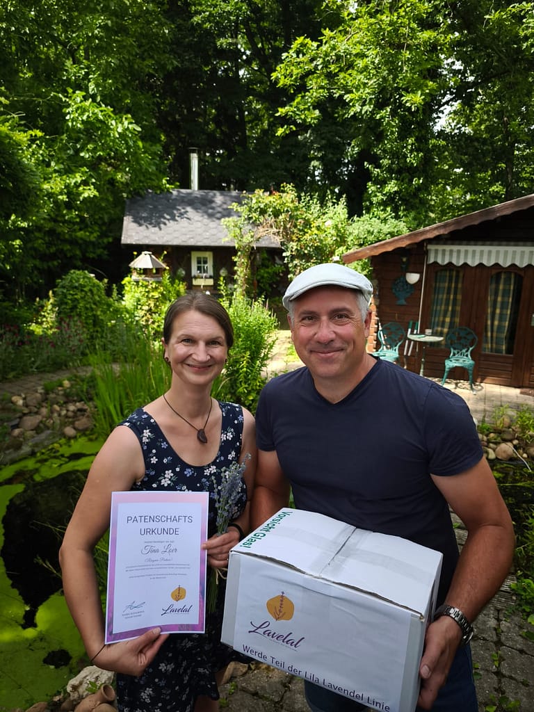 Gründer Mario Kohlmann überreicht Tina Loer, der ersten Patin bei LAVELAL, eine Geschenkbox und die offizielle Patenschaftsurkunde im Garten mit Lavendel und Teich.